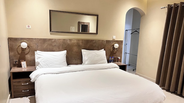 A hotel room with a large bed, a wooden headboard, a nightstand with a lamp, a mirror, and a view of a corridor. - Hamsun (Karachi)