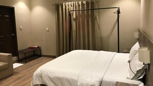 A hotel room with a bed, a bedside table, a chair, a curtain, and a wall-mounted air conditioning unit. - Hamsun (Karachi)