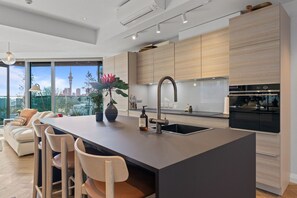 A modern kitchen with a central island, bar stools, and a large window overlooking a cityscape. - The Margot by Homello (Auckland)
