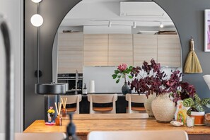 A modern kitchen with wooden cabinets, a dining area with a wooden table, and a large vase with flowers. - The Margot by Homello (Auckland)