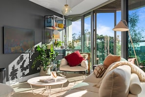A modern living room with a sofa, a coffee table, and a bookshelf. There is a large window providing a view of the outdoors. - The Margot by Homello (Auckland)