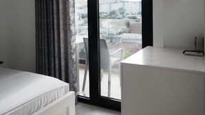 A modern bedroom with a bed, a desk, and a large window with a view of buildings. - Vila Mucodemi (Ksamil)