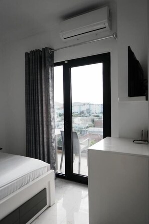 A modern bedroom with a bed, a desk, and a large window with a view of buildings. - Vila Mucodemi (Ksamil)