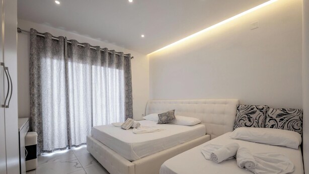 A hotel room with two beds, a bathroom, and a large window with curtains. - Vila Mucodemi (Ksamil)