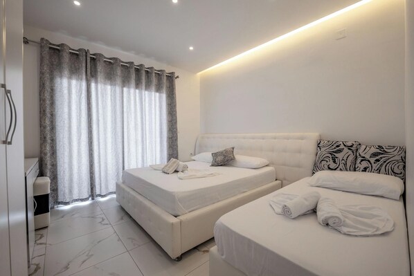 A hotel room with two beds, a bathroom, and a large window with curtains. - Vila Mucodemi (Ksamil)