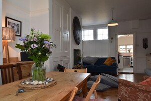 A dining area with a wooden table, chairs, a vase with flowers, and a view into the living room with a blue sofa.