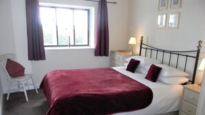 A neatly made bed with a red blanket, a bedside table with a lamp, a chair, and a window with curtains. - High Grange Cottages (Bedale)