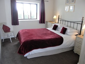 Cama bem arrumada com manta vermelha, mesa de cabeceira com abajur, cadeira e janela com cortinas.
