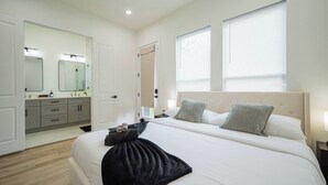 A hotel room with a bed, bedside tables, a bathroom with a mirror and sink, and two windows with blinds. - Modern 3BR | 3 Baths | Near NRG & Med Center (Houston)