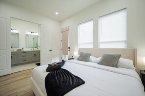 Una habitación de hotel con cama, mesitas de noche, baño con espejo y lavamanos, y dos ventanas con persianas.