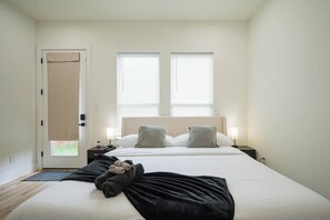 Un dormitorio con cama, dos mesitas de noche, una puerta con cortina y dos ventanas con persianas.
