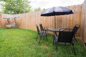 Un patio trasero con cerca de madera, una mesa redonda de vidrio, cuatro sillas negras y un paraguas azul.