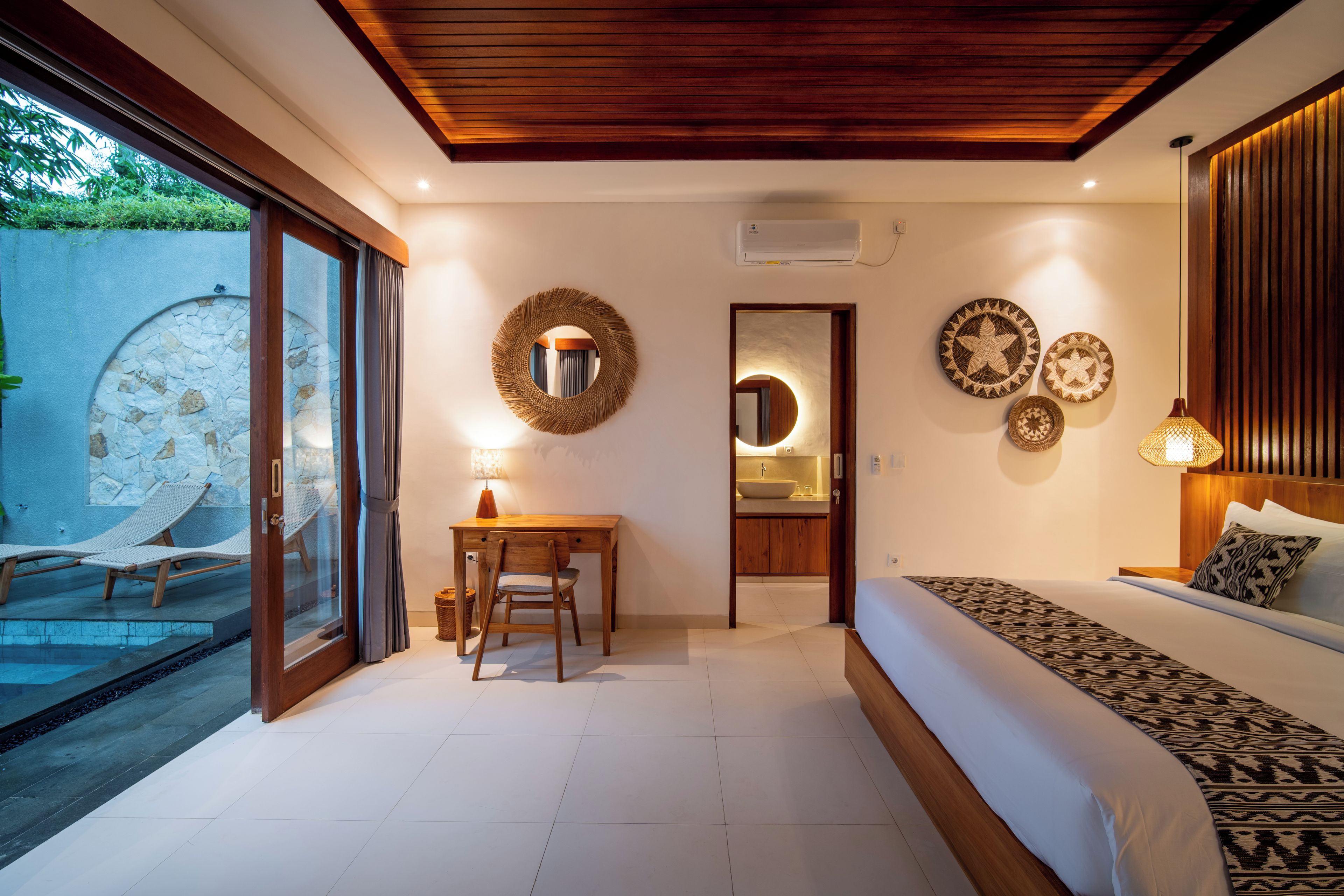 Un dormitorio con cama, escritorio y baño pequeño. Se observa una pared de piedra y una puerta de cristal que da a una zona de piscina.