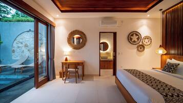 Un dormitorio con cama, escritorio y baño pequeño. Se observa una pared de piedra y una puerta de cristal que da a una zona de piscina.