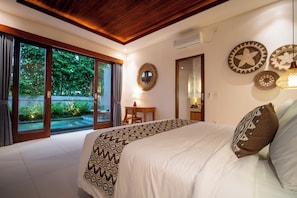 A bedroom with a large bed, a desk, and a view of a garden through glass doors. - Yoga Sri Ubud (Gianyar)
