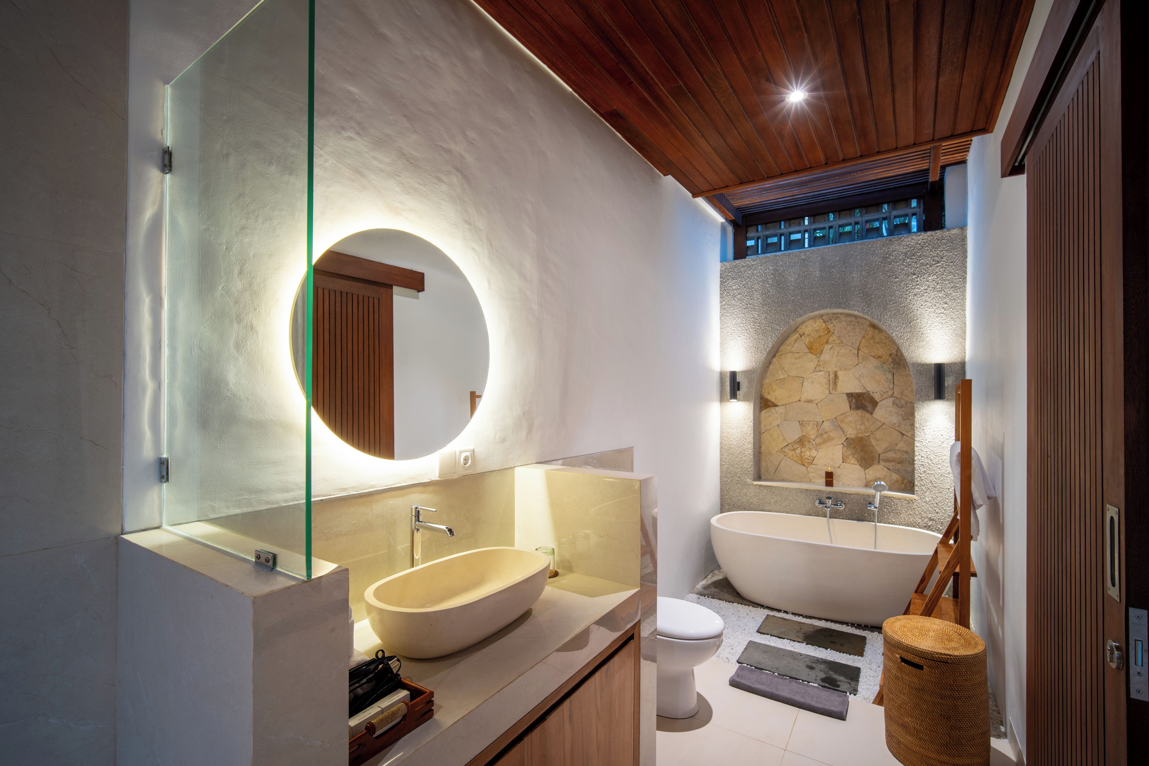 Un baño moderno con un lavamanos ovalado grande, una bañera independiente y un arco de pared de piedra.