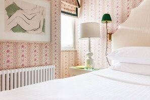 Ein Schlafzimmer mit einem Bett, einem Heizkörper, einer Wandleuchte, einem gerahmten Kunstwerk und einem Fenster mit floralem Vorhang.