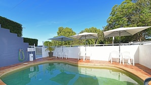 Una piscina con borde de ladrillo, rodeada de sillas y sombrillas blancas, y una pared blanca con una reja.