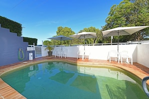A swimming pool with a brick border, surrounded by white chairs and umbrellas, and a white wall with a gate. - Belvedere Apartments (Sunshine Coast)