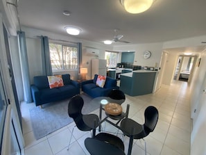 A modern living room with a sofa set, a dining table with chairs, a kitchen area, and a ceiling fan. - Belvedere Apartments (Sunshine Coast)