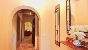 Un pasillo con una entrada arqueada que conduce a una habitación, un espejo con marco decorativo y un jarrón con flores sobre una estantería.