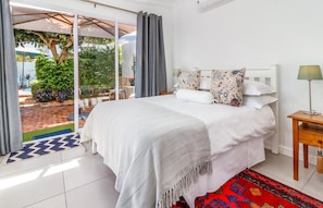 A bedroom with a bed, a nightstand, a lamp, and a view of the garden through a sliding door. - Bleu Cobalt Cottage (Franschhoek)
