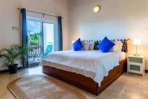 Quarto com uma cama grande, mesa de cabeceira, abajur, planta em vaso e vista para o exterior.