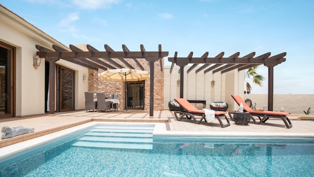 Een zwembad met ligbedden en een pergola, een stenen muur en uitzicht op het strand.