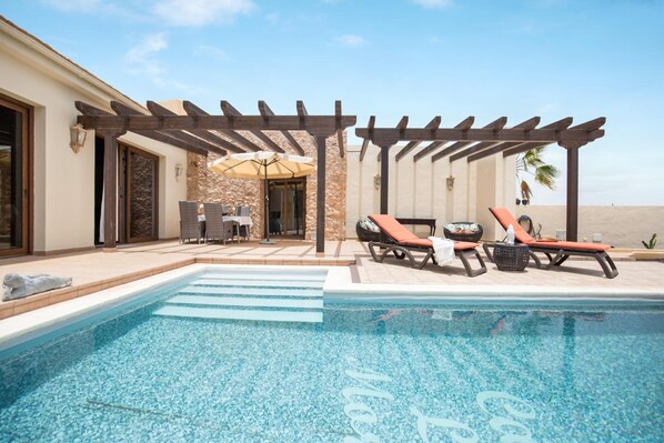 A swimming pool with lounge chairs and a pergola, a stone wall, and a view of the beach. - Casa Rural La Morisca (Tuineje)
