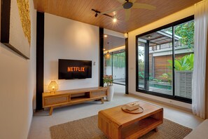 Ein modernes Wohnzimmer mit einem Flachbildfernseher, Holzmöbeln und Blick auf einen Garten durch große Schiebetüren.