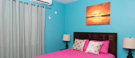 Un dormitorio con cama, cortinas, aire acondicionado, un cuadro y lámparas de noche.