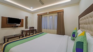 Quarto de hotel com cama, escrivaninha, cadeira, televisão e janela com cortinas.