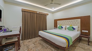 Quarto de hotel com cama, escrivaninha, cadeira, ventoinha no teto e janela com cortinas.