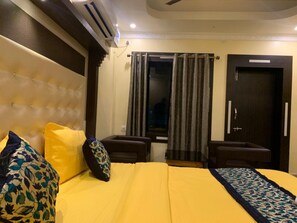 Ein Hotelzimmer mit einem gebogenem Kopfende, einem Bett mit gelber Bettwäsche, zwei Sesseln, einem kleinen Tisch und einer dunklen Tür.