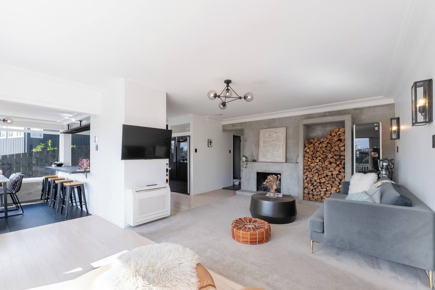 A modern living room with a grey sofa, a black coffee table, and a fireplace. There is a dining area with a table and chairs, and a kitchen with a refrigerator and a TV.