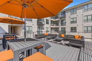 An outdoor deck with seating and umbrellas, a modern building with balconies in the background. - Heart of The Domain  1BR w/ Pool, Gym & Free Parking (Austin)