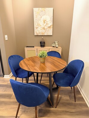 A dining area with a round wooden table surrounded by four blue chairs, a decorative Buddha head, a scooter, and a framed floral artwork on the wall. - Heart of The Domain  1BR w/ Pool, Gym & Free Parking (Austin)