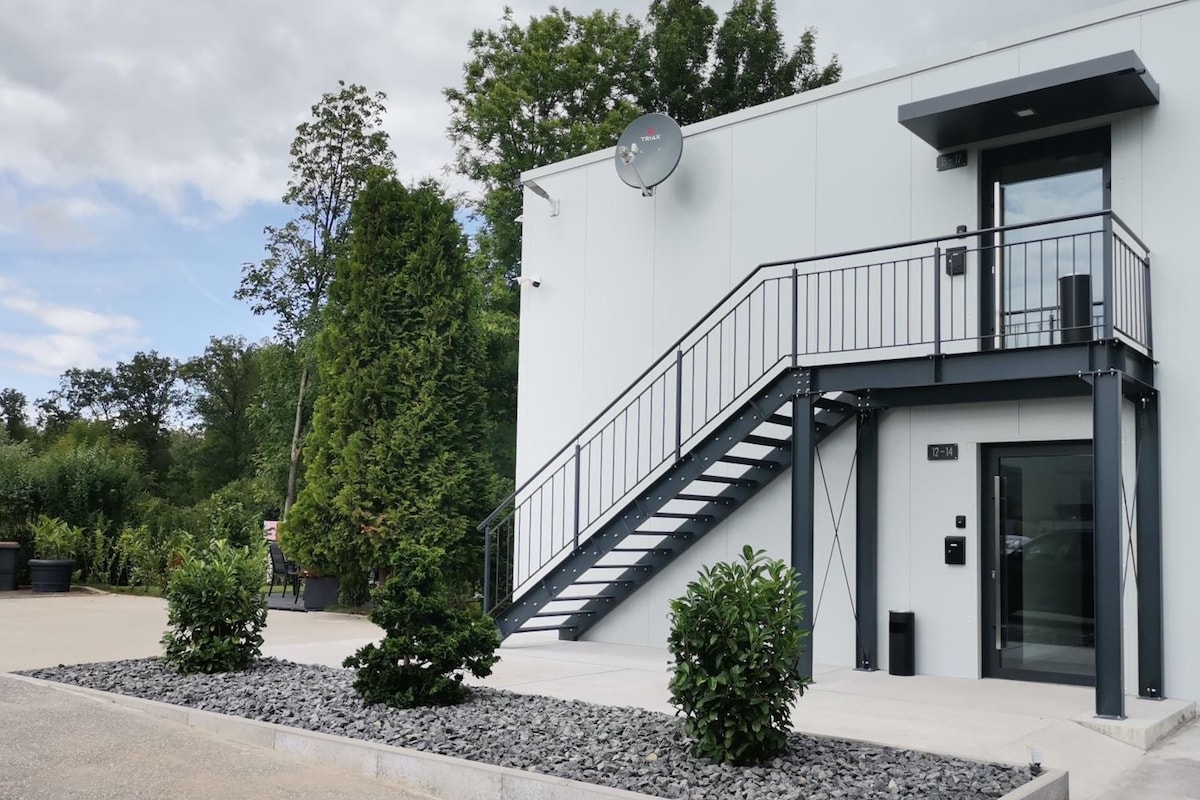 Edificio moderno con fachada blanca, antena parabólica, escalera que conduce a un balcón y un pequeño jardín con arbustos y grava.