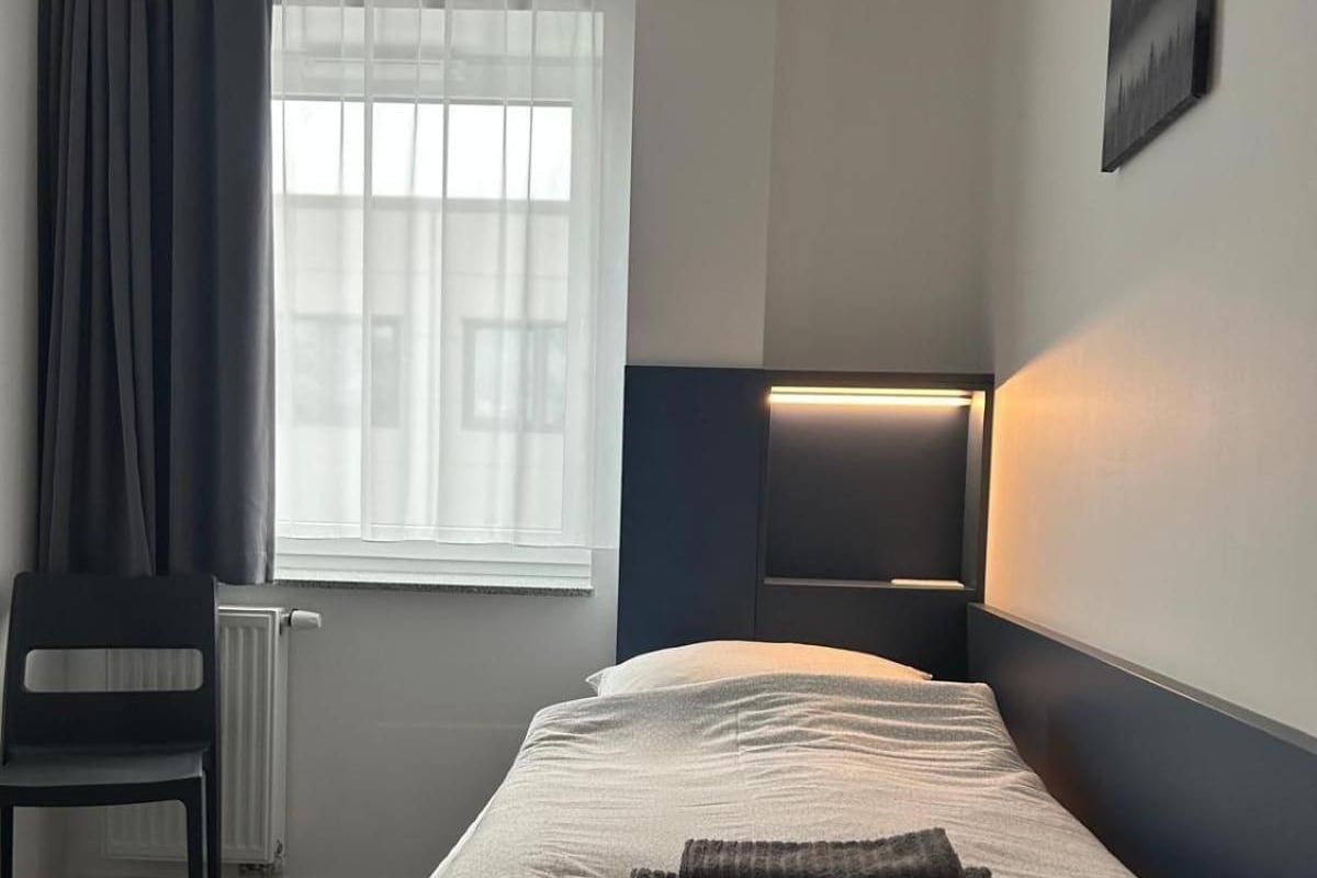 Una cama individual con cabecera, una silla, un radiador, una ventana con cortinas e iluminación de pared.