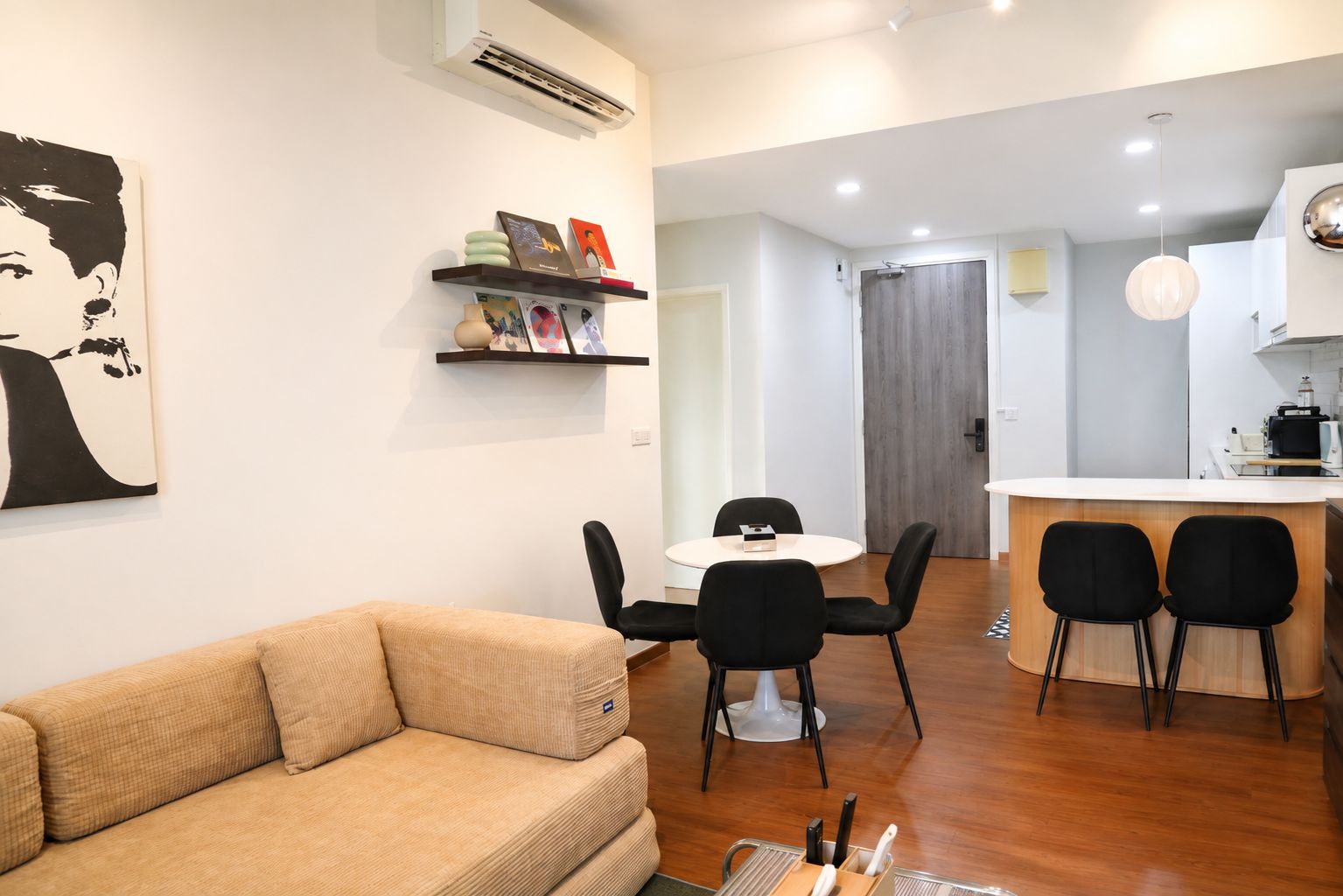 A modern living room with a sofa, a dining table with chairs, a kitchen area with a counter and chairs, and a hallway leading to a door.