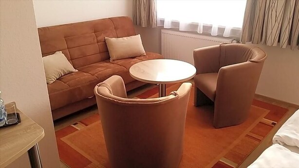 Et værelse med en brun sofa, to lænestole, et lille rundt bord og en seng med hvide sengetøj.