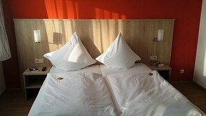 En dobbeltseng med hvide sengetøj og to puder, et trægavl, natborde med lamper, og en rød væg.