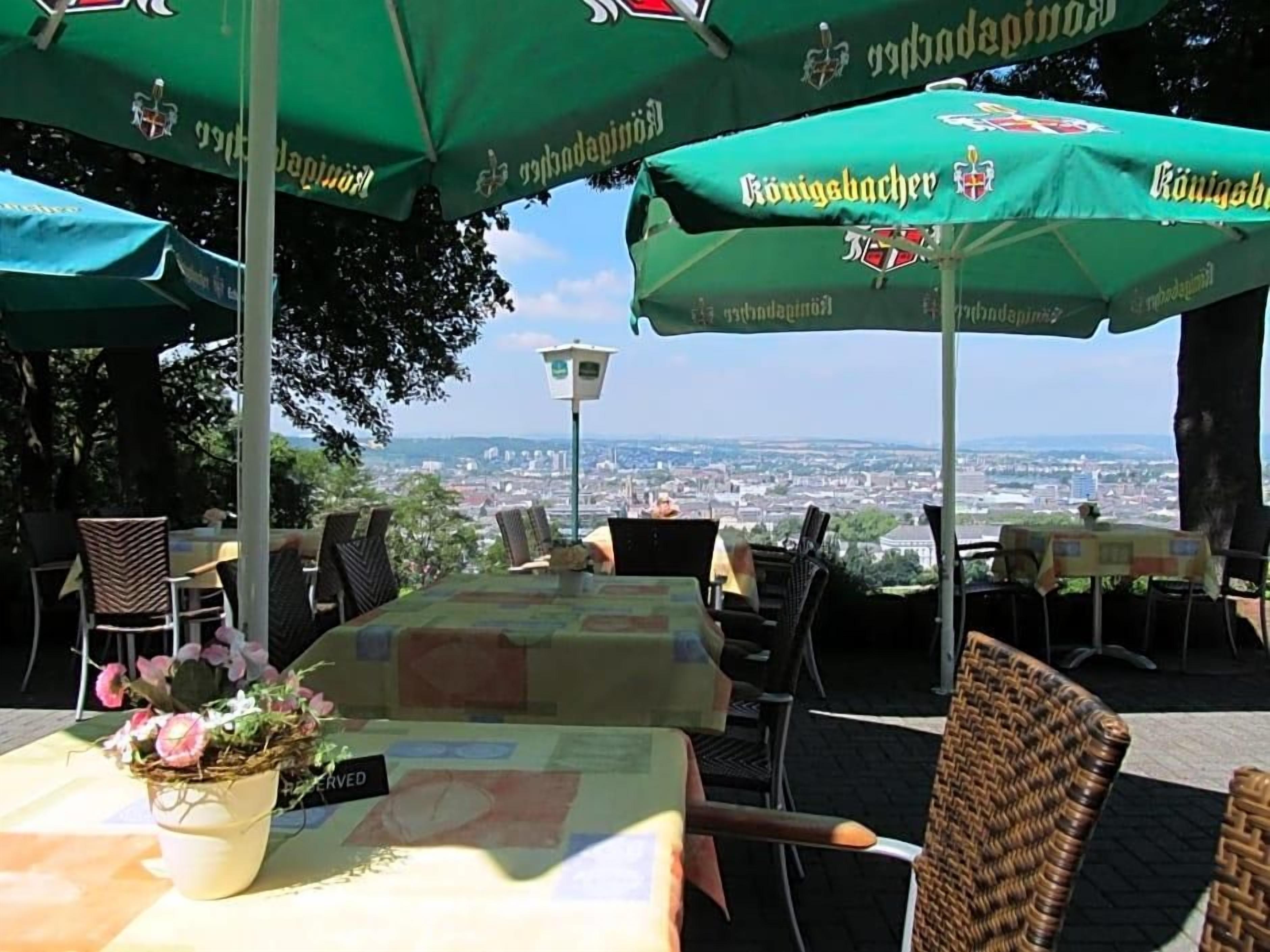 Ein Außenbereich mit Tischen und Stühlen, grünen Sonnenschirmen und Blick auf eine Stadtlandschaft.