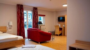 Et hotelværelse med seng, rød sofa, lille bord, lampe, fjernsyn og et vindue med gardiner.