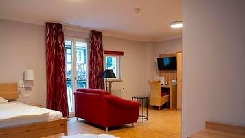 Ein Hotelzimmer mit einem Bett, einem roten Schlafsofa, einem kleinen Tisch, einer Lampe, einem Fernseher und einem Fenster mit Vorhängen.