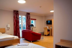 Quarto de hotel com cama, sofá vermelho, pequena mesa, abajur, televisão e janela com cortinas.
