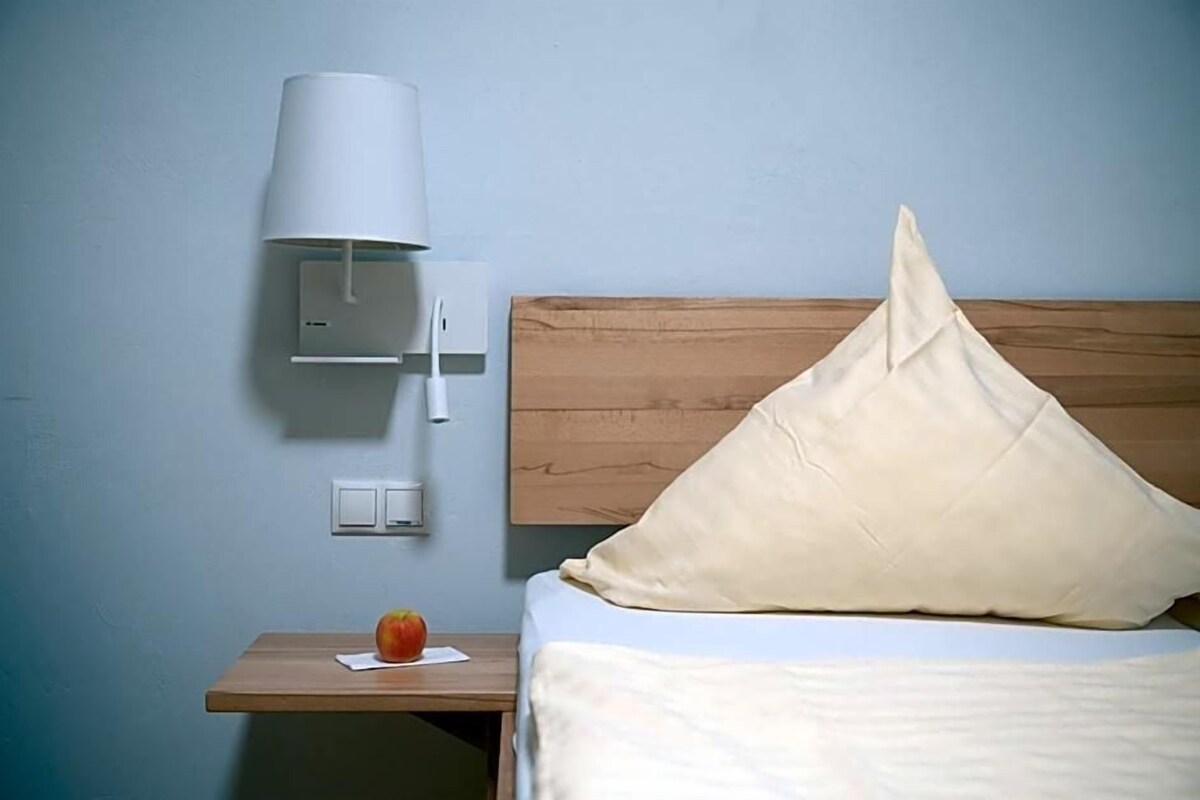 A hotel room with a wooden headboard, a white pillow, a bedside table with a lamp, and a fruit on the table.