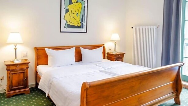 A bedroom with a wooden bed, two bedside tables with lamps, a radiator, and a window with curtains. - Brauhaus In Spandau (Berlin)