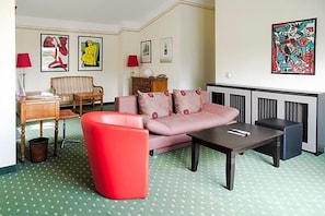 Ein Zimmer mit einer Couch, einem roten Sessel, einem Tisch, einer Lampe und Kunstwerken an den Wänden.