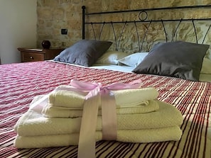 A neatly made bed with a striped bedspread, a stack of folded towels with a ribbon, and a stone wall in the background. - Masseria Monte Pizzi (Carovilli)